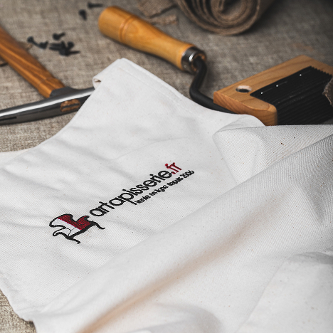 Tablier blanc brodé du logo artapisserie.fr, posé sur des outils de tapisserie à manche en bois